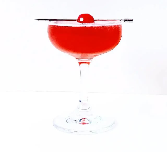 Elegant red cocktail in a coupe glass, garnished with a maraschino cherry on a metal pick.