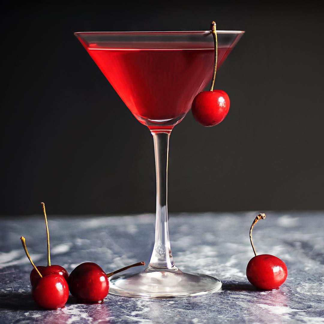 Red cocktail in a martini glass with a cherry garnish on a marble surface, surrounded by fresh cherries.