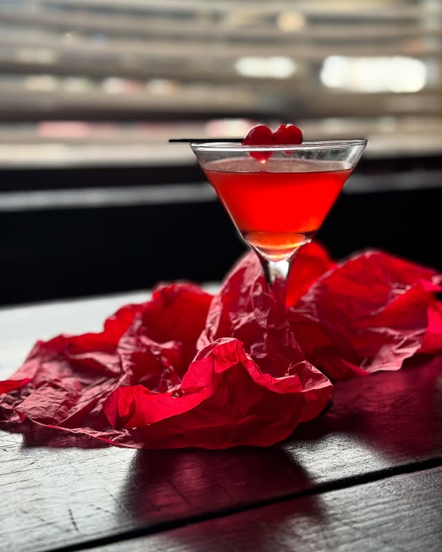 Red cocktail in a martini glass, garnished with cherries, on a table with red fabric, against a blurred background.