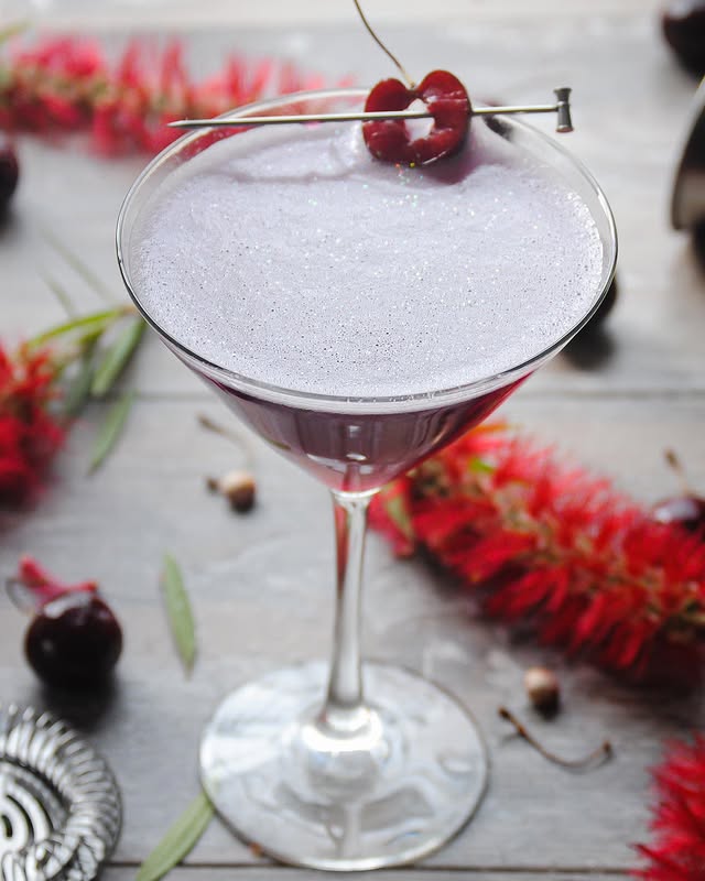 Purple cocktail garnished with cherries in a martini glass, surrounded by red flowers on a rustic table.