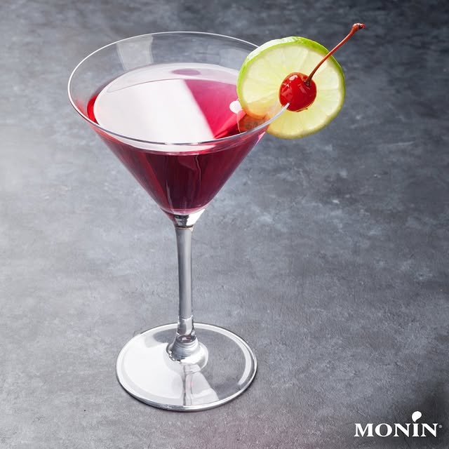 Red cocktail in a martini glass garnished with a lime slice and cherry, on a dark surface. Monin logo in corner.