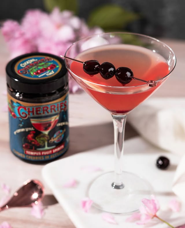 Elegant pink cocktail garnished with cherries, next to a jar of gourmet cherries on a table with floral decor.