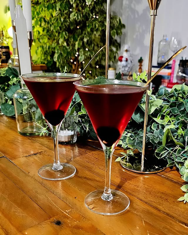 Two elegant red cocktails on a wooden table with a lush green backdrop.