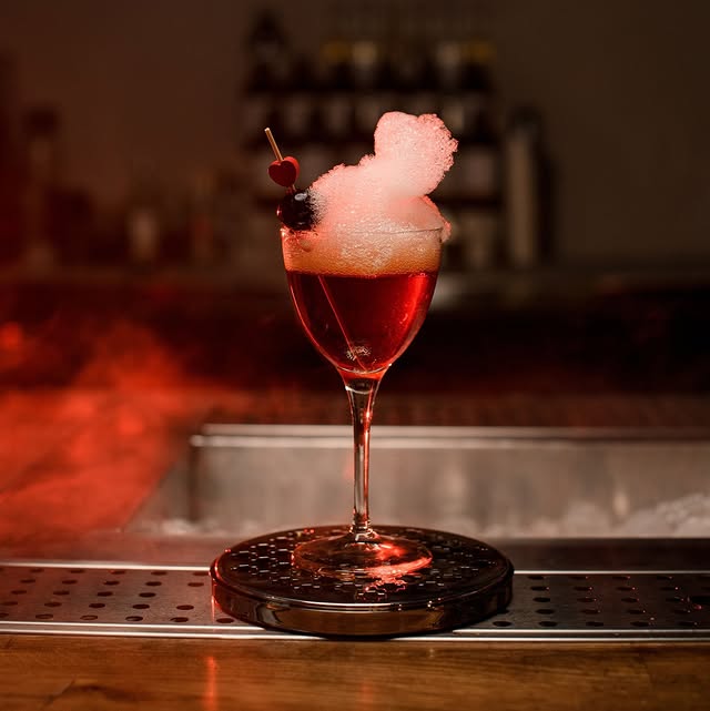 Elegant cocktail with foam, cherry, and blueberry garnish, set on a bar with ambient red lighting.