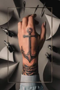 Hand with anchor tattoo, surrounded by tattoo ink bottles and tools on a gray background.