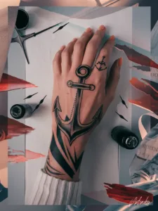 Hand with detailed anchor tattoo on wrist and finger, surrounded by art supplies on a creative workspace.