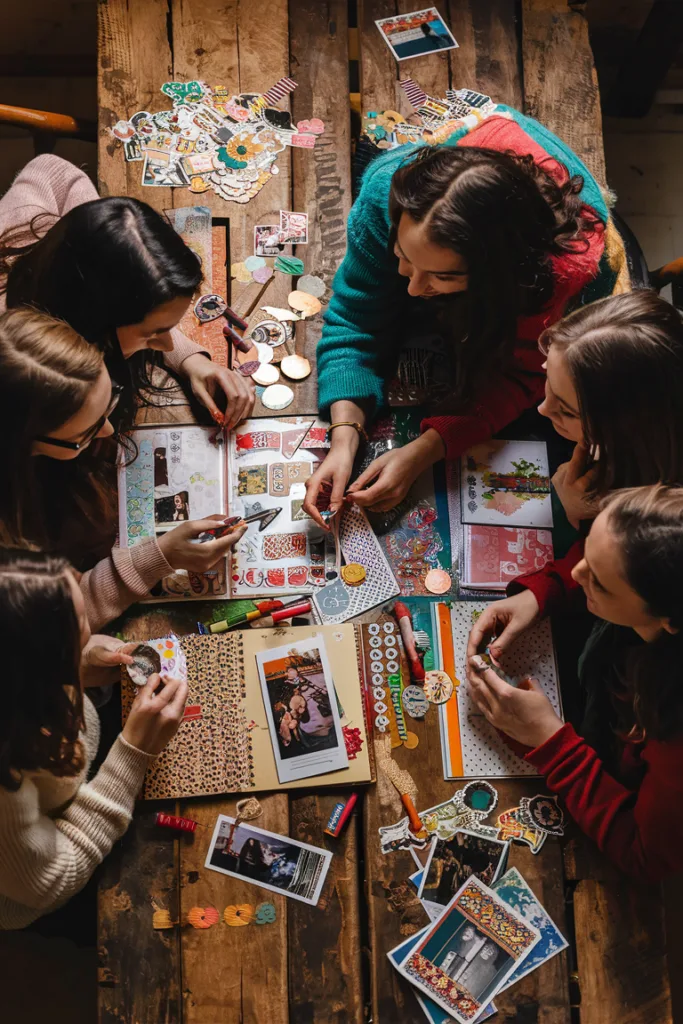 Group of friends crafting colorful scrapbook pages on a wooden table, surrounded by photos and decorations.