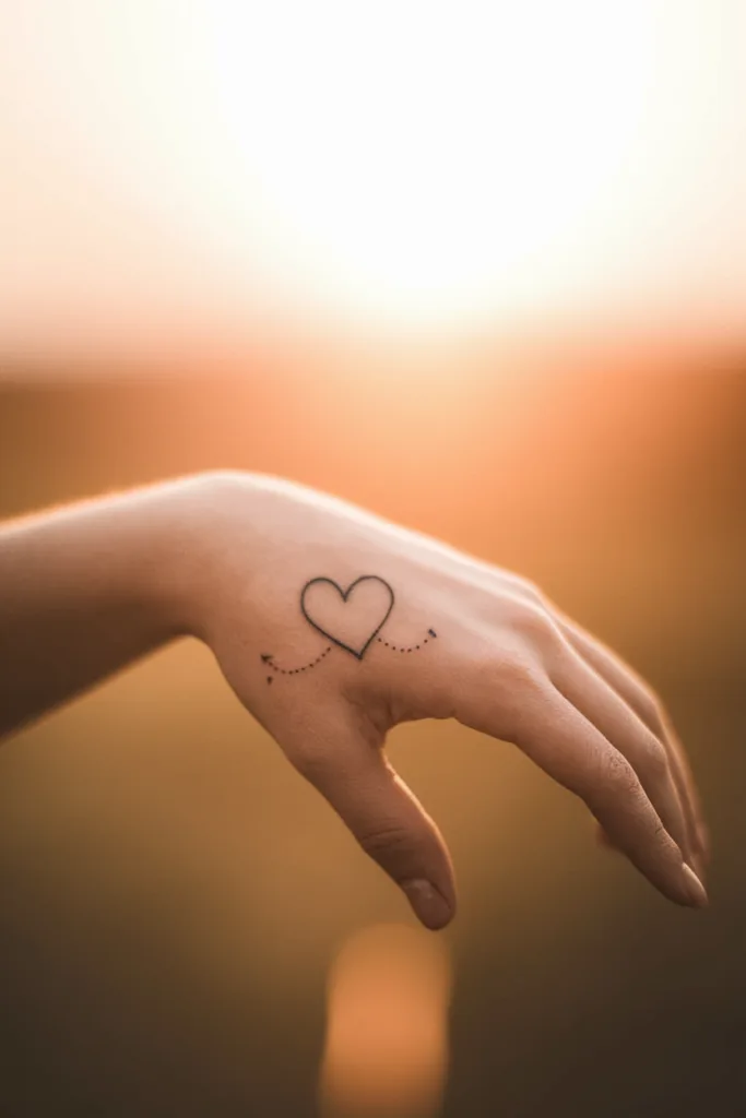 Minimalist heart tattoo on a hand against a warm, blurred sunset background.