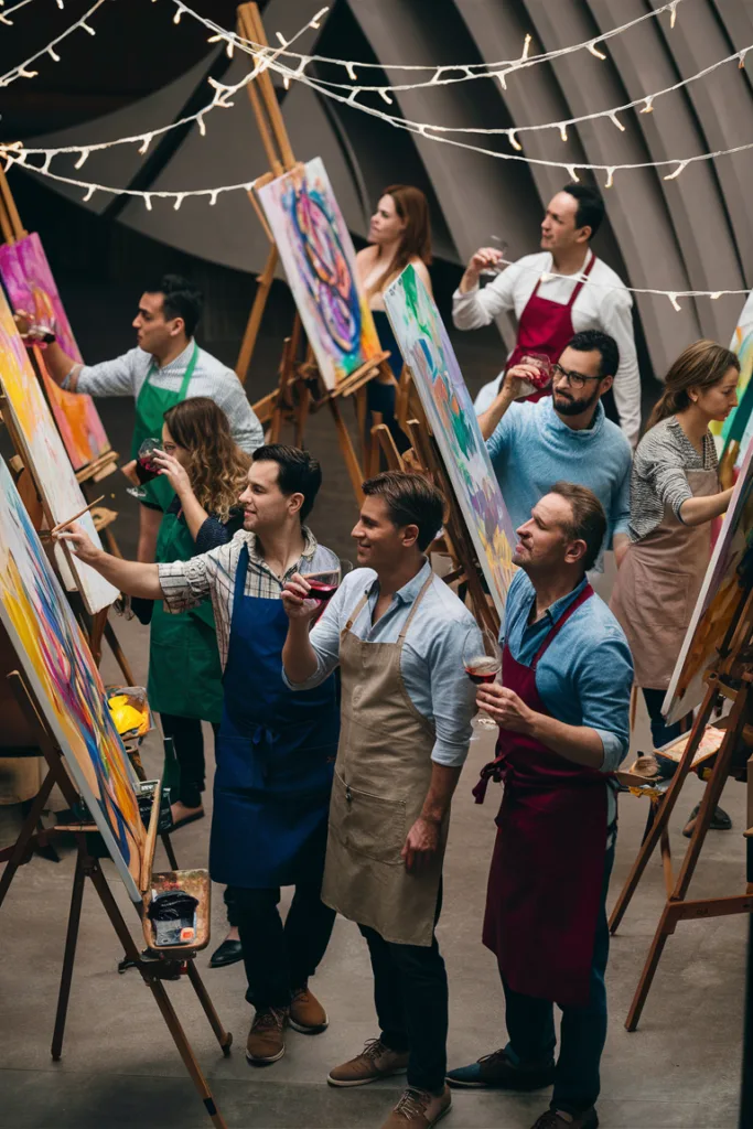 Group enjoying a wine and painting class, creating colorful artworks on easels, under decorative string lights.