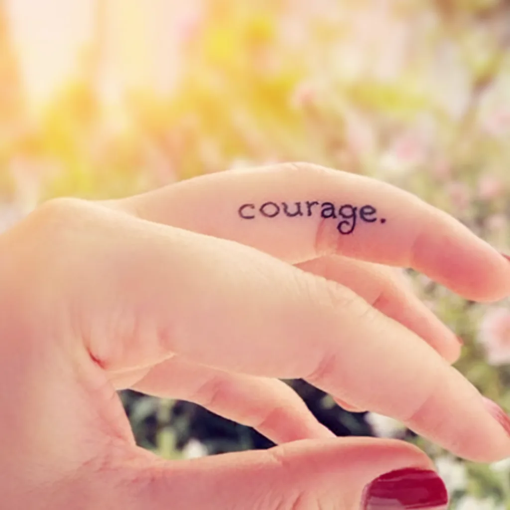 Close-up of a finger tattoo with the word courage in an outdoor setting with sunlight and flowers in the background.